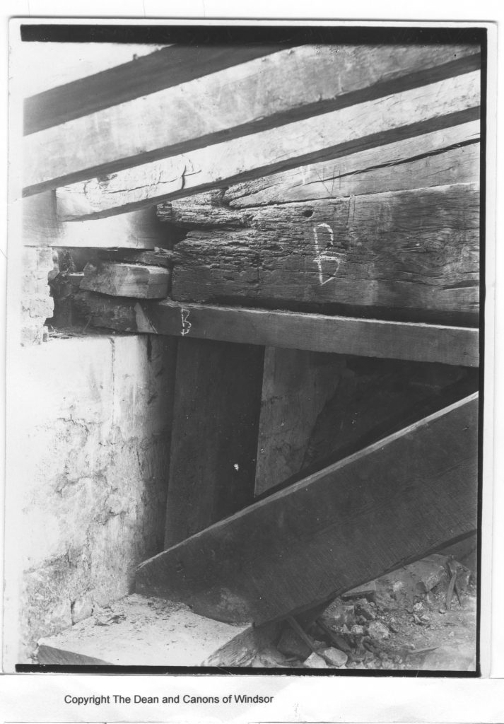 A black and white photograph showing dark wooden beams forming a triangular shape, braced against a stone wall. The end of one beam (labelled 'B' in chalk) is badly decayed, leaving a gap between it and the wall which has been filled with smaller pieces of timber.