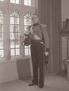 A black and white photograph depicting a middle-aged white man with short, dark hair and round glasses wearing the uniform of the Military Knights of Windsor. He is wearing eith medals on his left breast and his hat is tucked under his right arm. He is standing in a furnished living room in front of large, mullioned windows.