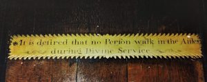 A brass plate on a wooden background reading 'It is desired that no Person walk in the Aisle during Divine Service.' The edges of the plate are scalloped and there are decorative curls next to the text.