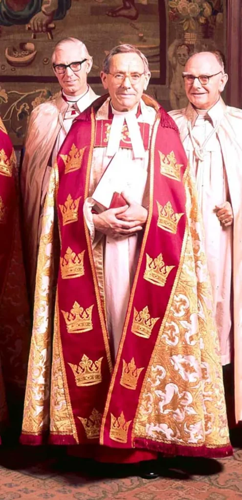 Portrait photograph of a white man with short, grey hair and round glasses, wearing a cassock, clerical bands and a red and gold cope.