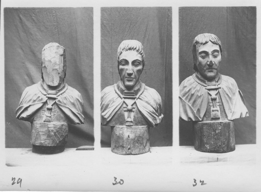 Three carved busts of men in cloaks, two of them have parts of their face cut off. Photograph is in black and white, background is plain.